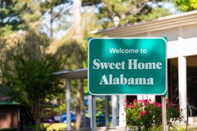 Green roadside sign reading Welcome to Sweet Home Alabama near a building and trees