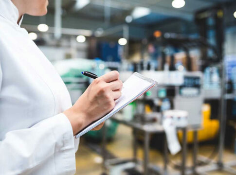 Person in lab coat writing on clipboard in factory.