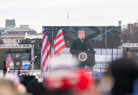 A large screen displays a speaker at a political rally with American flags in the foreground